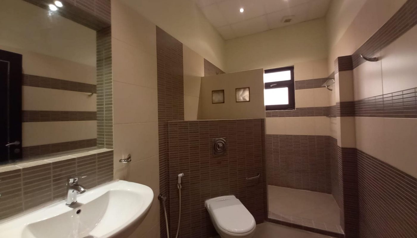 A modern bathroom with a white sink, a wall-mounted toilet, and a shower area separated by a tiled partition. The walls are tiled in brown and beige.