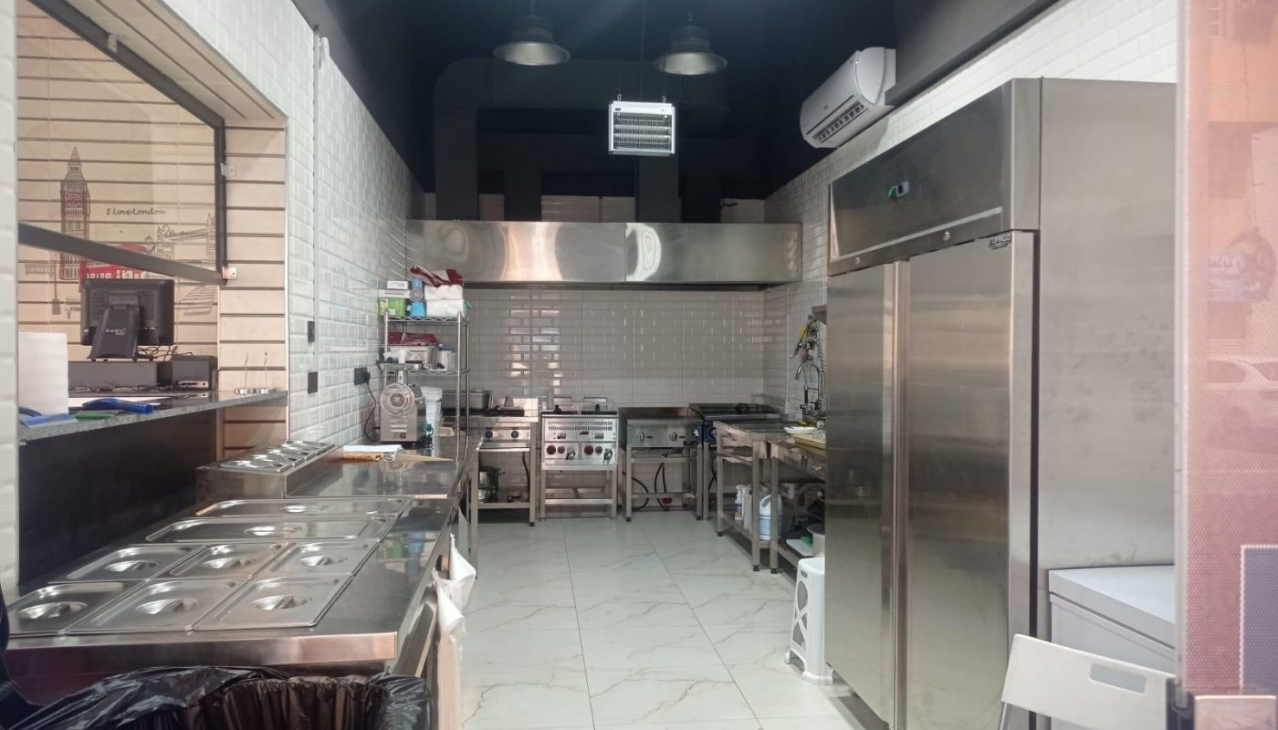 Commercial kitchen with stainless steel appliances, a large fridge, food prep stations, tiled walls, and overhead lighting. The area is clean and organized.