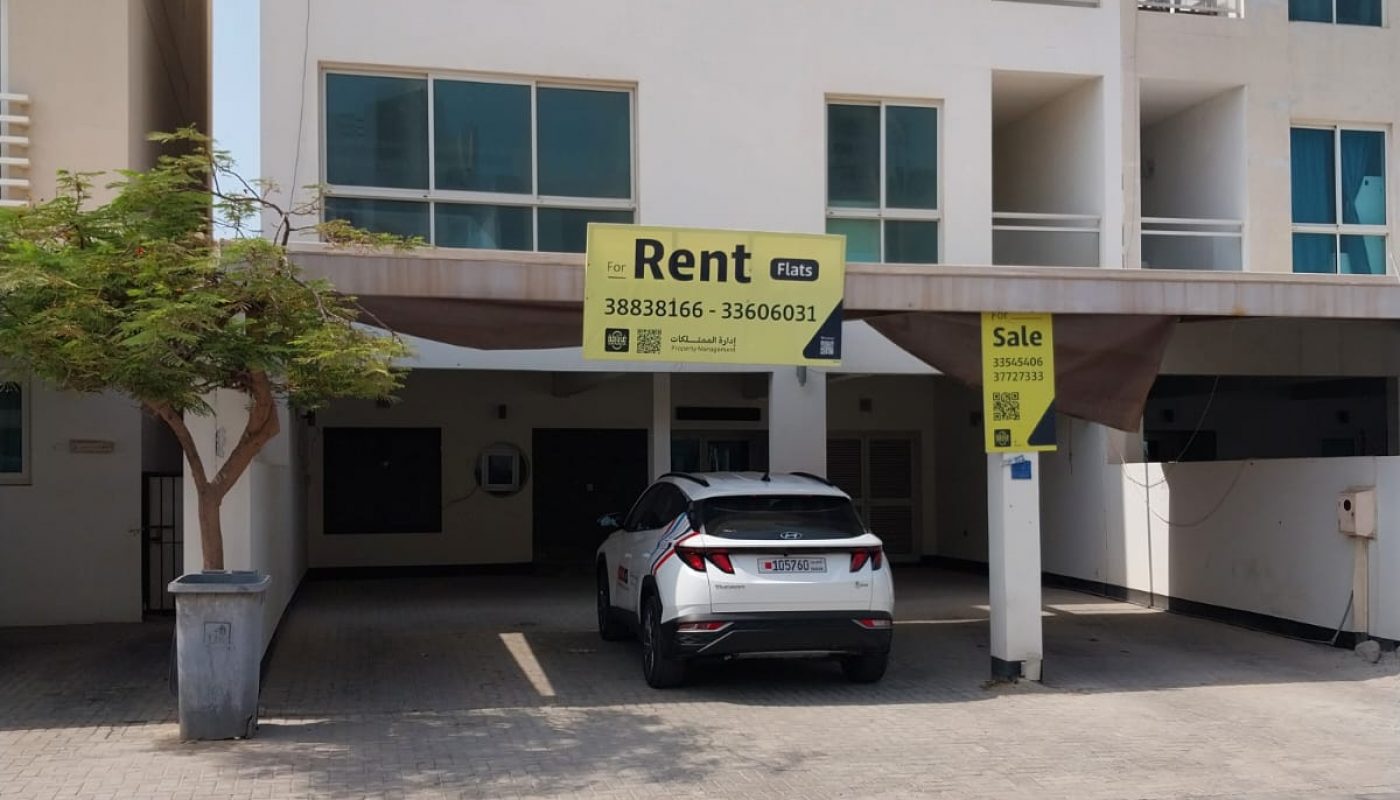 A white, modern three-story building with a car parked in front. Signs indicate the property is available for rent and for sale.