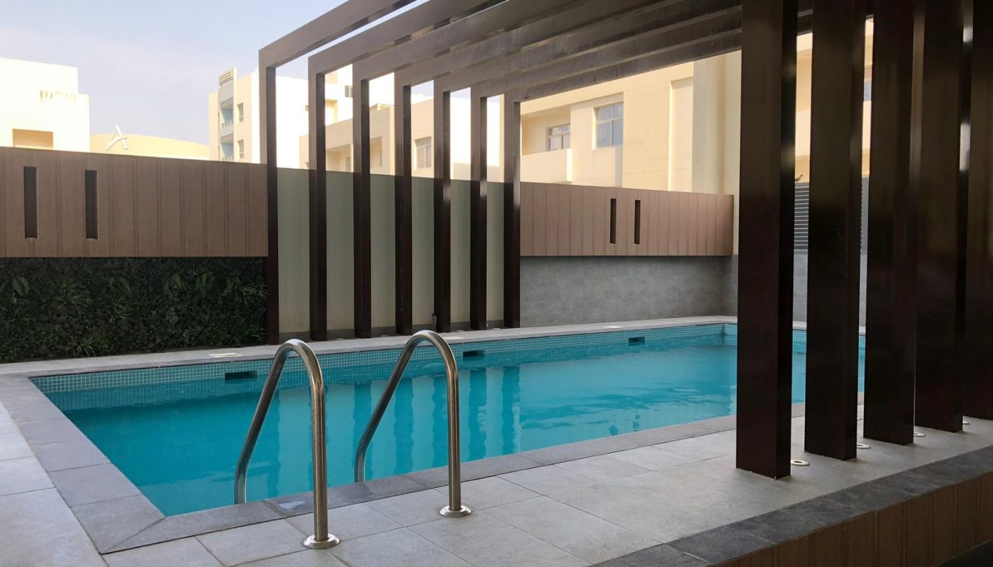 Rectangular indoor swimming pool with metal handrails, surrounded by tiled flooring and vertical wooden beams, next to a wall with a green panel. Buildings are visible in the background.