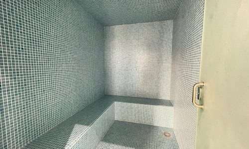 A small steam room with blue mosaic tiled walls, ceiling, and bench seating, and a door with a handle is just one of the luxurious amenities offered in this AMWAJ flat for sale.