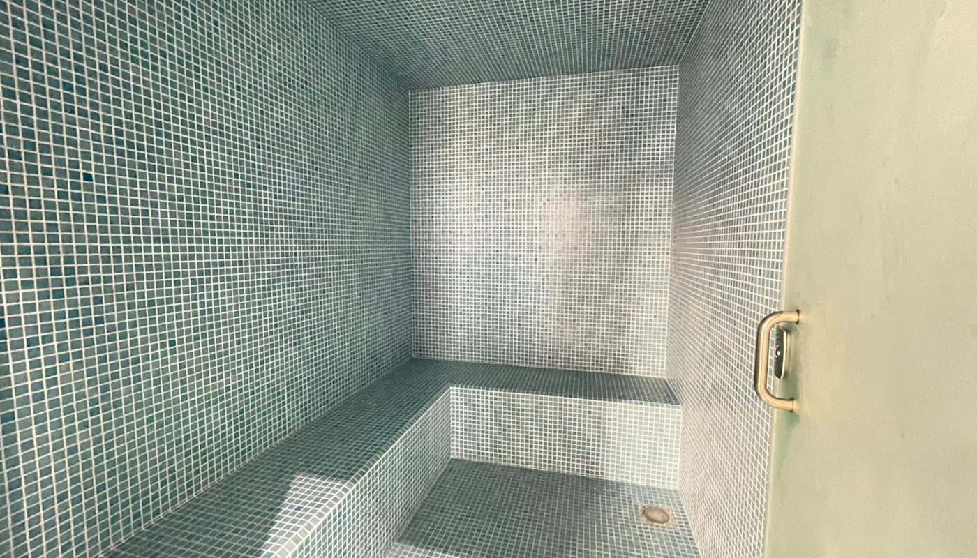 A tiled steam room with a built-in bench, light blue mosaic tiles, a ceiling light, and a door handle visible on the right.