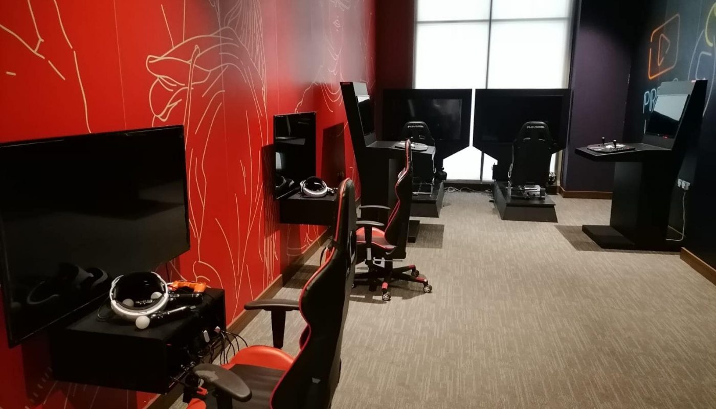 A room with gaming chairs, steering wheels, and monitors, set up for simulation gaming along red and black walls.