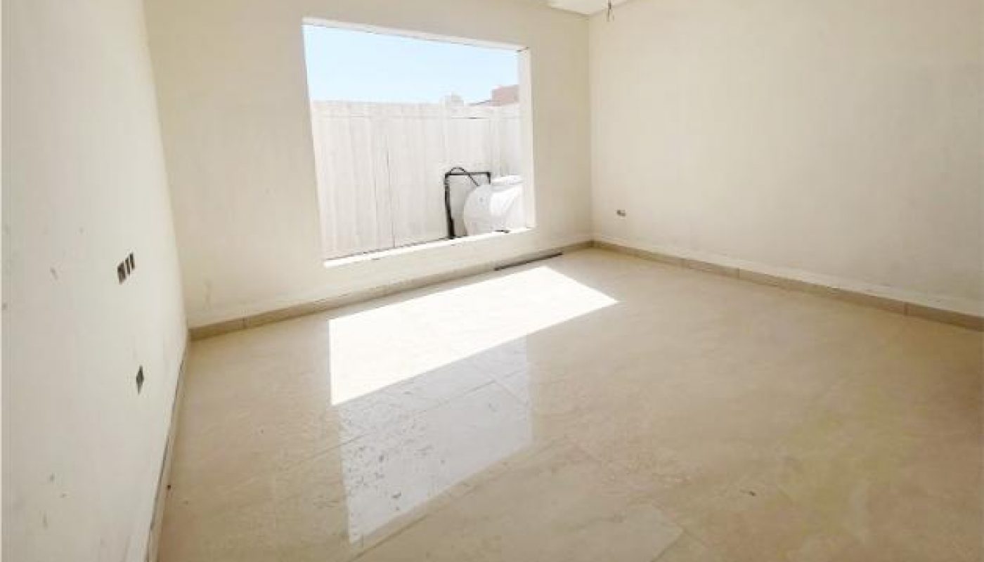 Empty, unfurnished room with cream-colored walls and floor, a large window letting in sunlight, and exposed ceiling light fixtures.