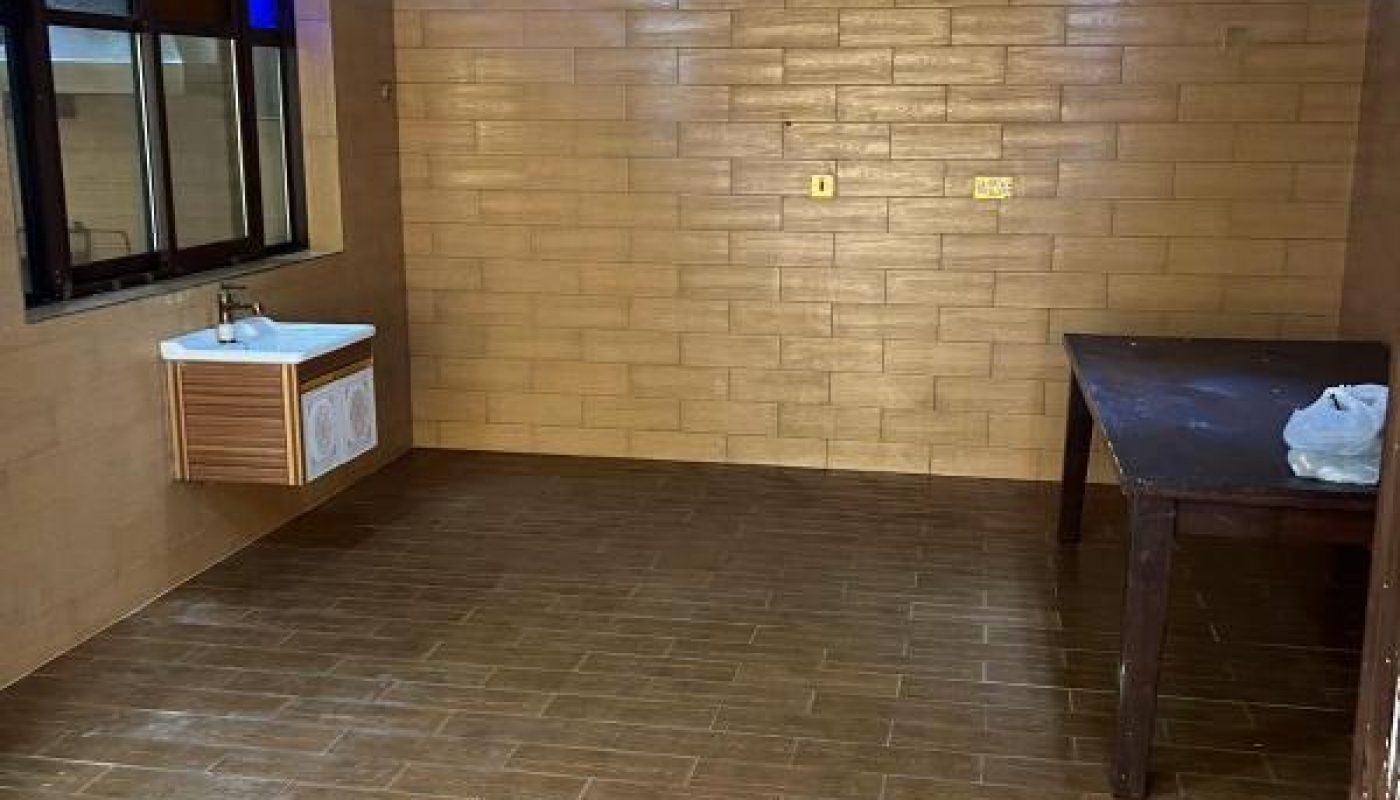 An empty room with brown tile floors and walls, a sink with a cabinet on the left, a large wooden table on the right, and three hanging pendant lights from the ceiling.