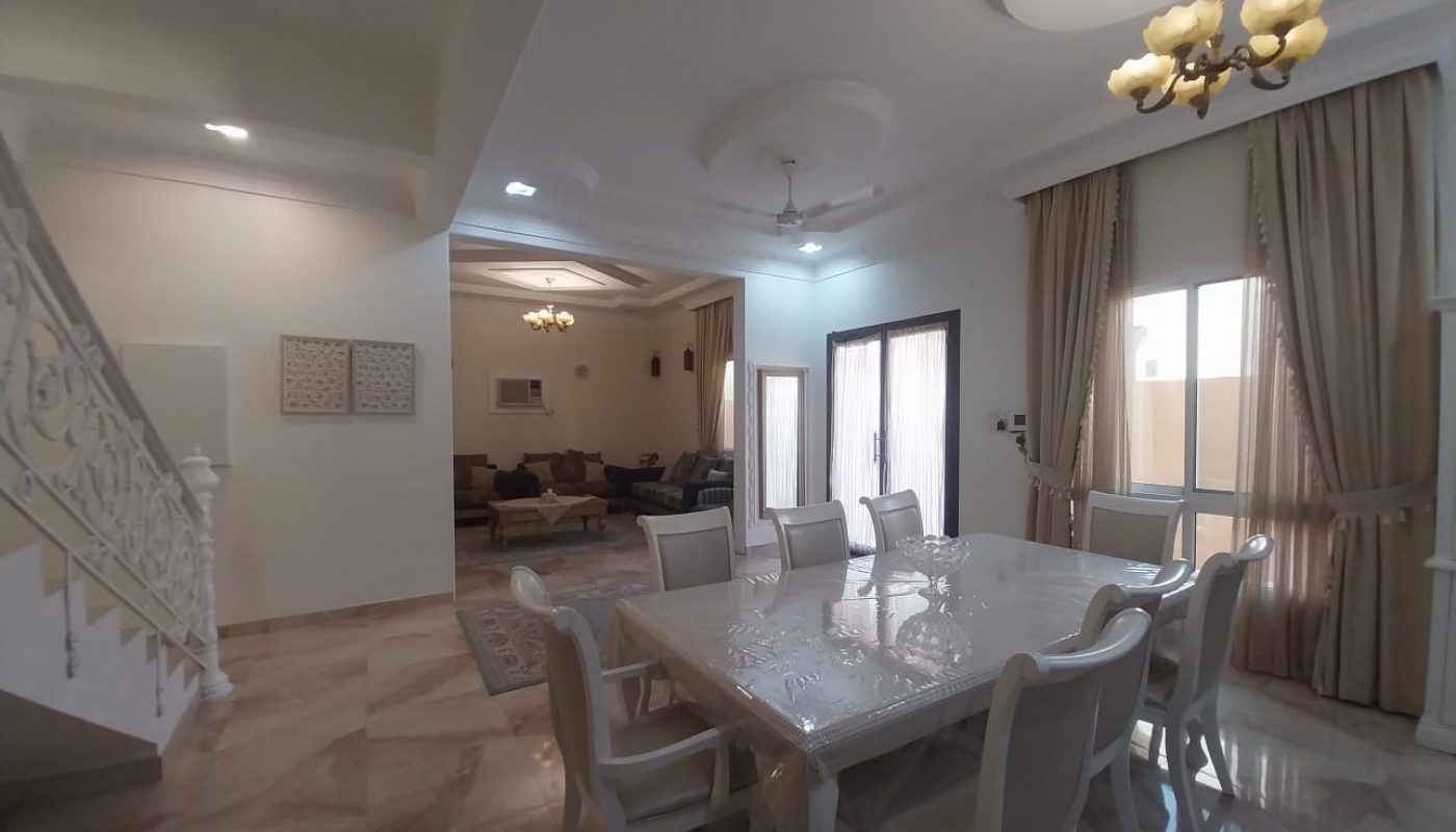 A spacious dining area with a white table and chairs, adjacent to a living room, staircase, and large windows with beige curtains. Marble flooring and chandelier lighting are visible.