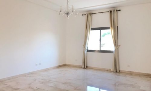 Empty living room with shiny tiled floor, white walls, and a window dressed with beige curtains under a chandelier.