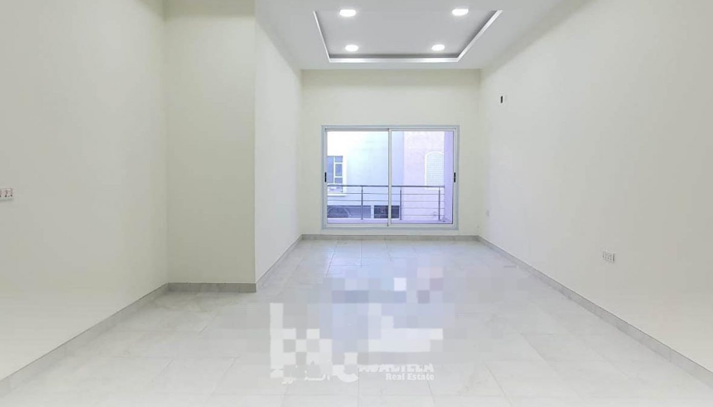 Empty room with white walls, recessed ceiling lights, and large window at the far end. The floor is tiled, and there is a balcony visible through the window.