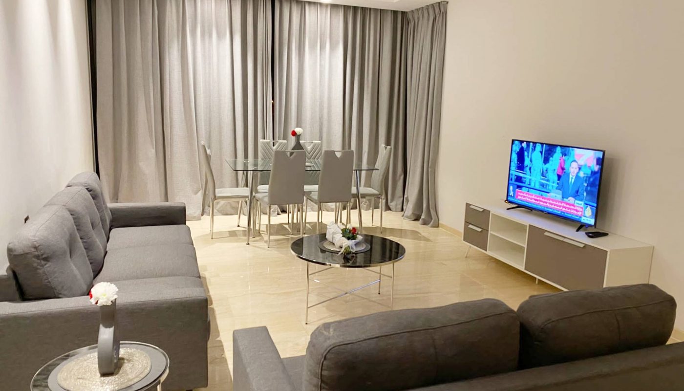 A fully furnished living room in an apartment for rent, featuring a grey couch and TV, located in the Dilmunia area.
