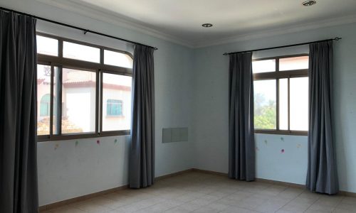 Empty room with two large windows covered by gray curtains. The walls are light blue with faint marks and the floor is tiled. Ceiling lights are also visible.