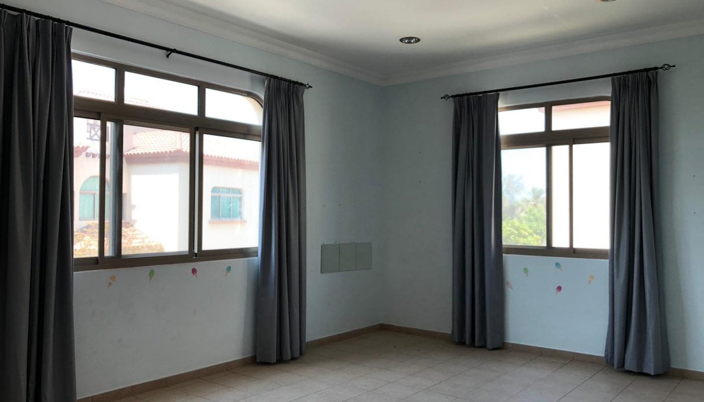 Empty room with two large windows covered by gray curtains. The walls are light blue with faint marks and the floor is tiled. Ceiling lights are also visible.