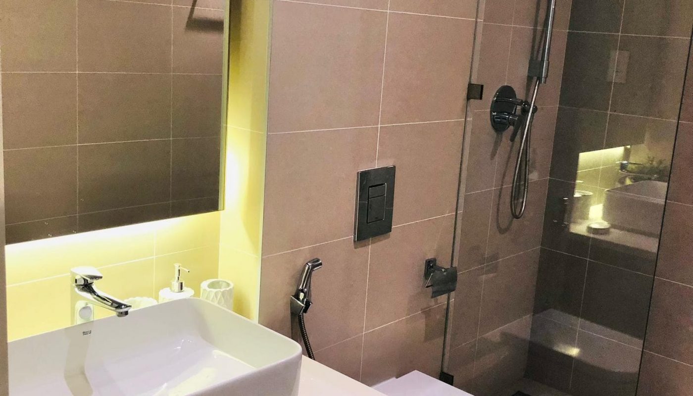 A modern bathroom in a 1-bedroom flat for sale at Marassi Boulevard, featuring a square sink, a wall-mounted toilet, and a glass-enclosed shower. The walls and fixtures are in neutral tones, and a large mirror is illuminated by built-in lighting.