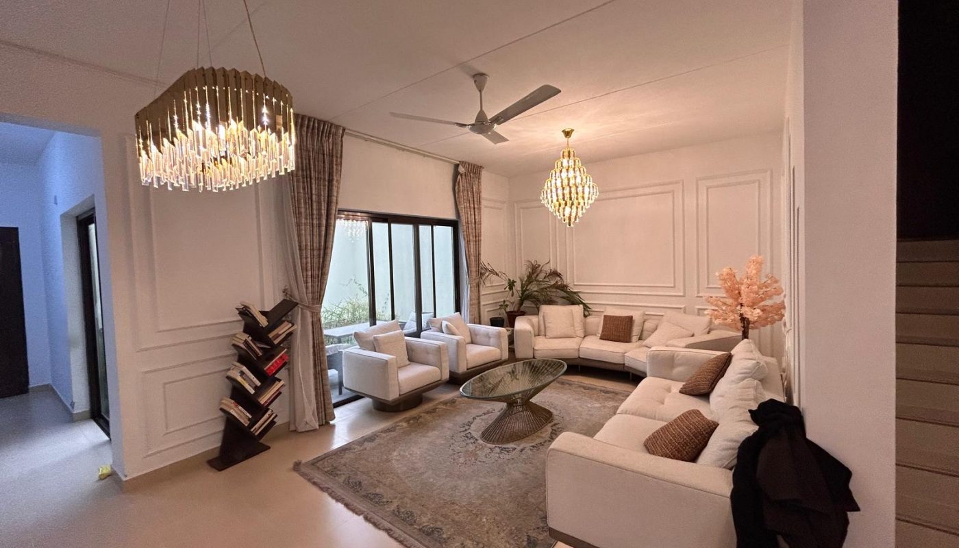 A modern living room with white sofas, a glass coffee table, two chandeliers, large windows, neutral walls, and a bookshelf near the entrance.