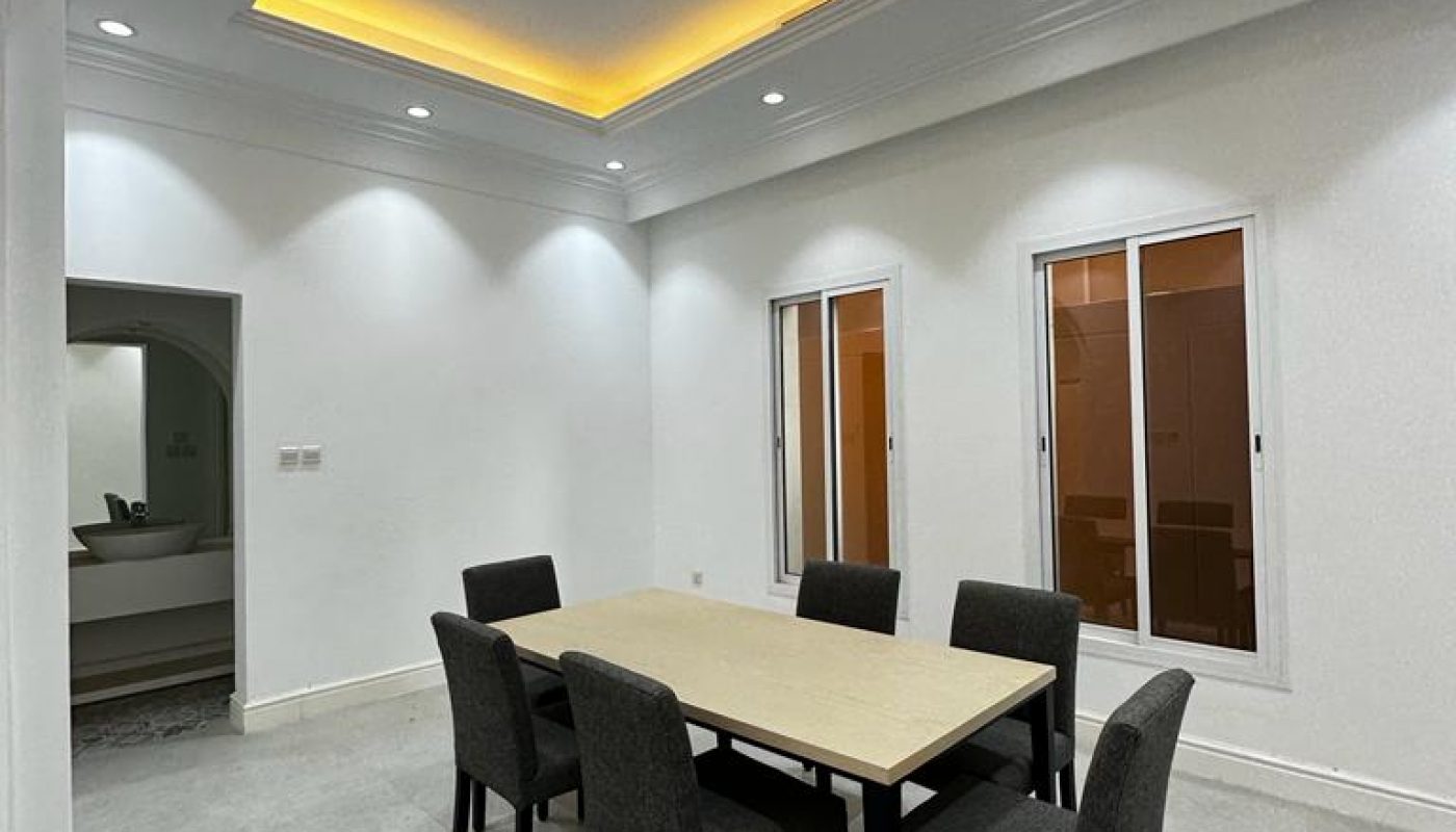 A modern dining room with a rectangular table, six gray chairs, white walls, large windows, and a ceiling with recessed lighting.