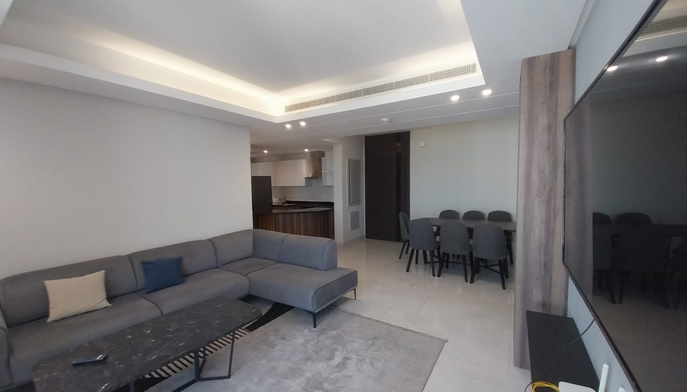 Modern living room with a gray sectional sofa, black coffee table, and dining area with a table and eight chairs; television and router visible on the right.
