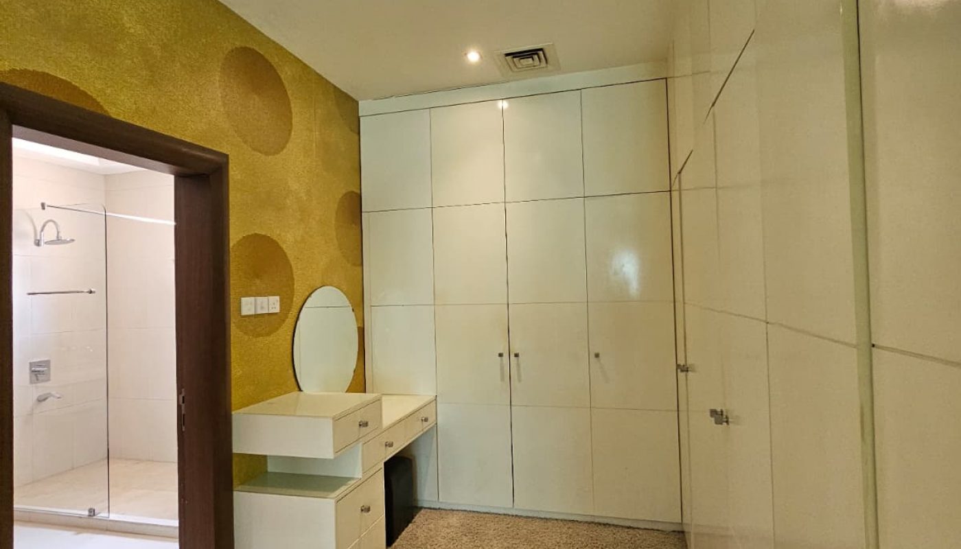 A narrow room with white built-in cabinets, a small desk with a mirror, beige carpet, and a door leading to a bathroom.