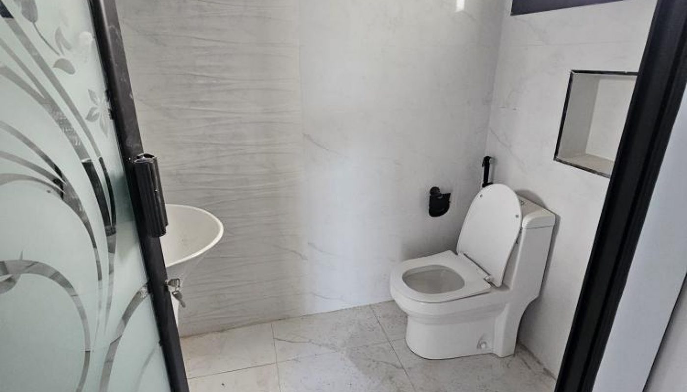 A small bathroom with a white toilet, a wall-mounted sink, a towel ring, and frosted glass door with a floral pattern. The walls and floor are white tile.