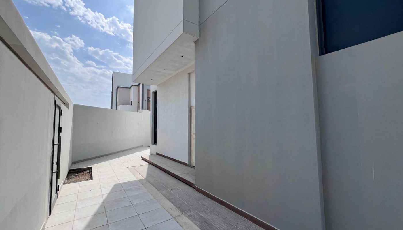 Narrow outdoor walkway beside a modern building with light gray walls and tiled flooring, under a partly cloudy sky.