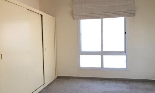 A bright, empty room with beige walls, a light gray carpet, a large window with a white curtain, and a closed white wardrobe.