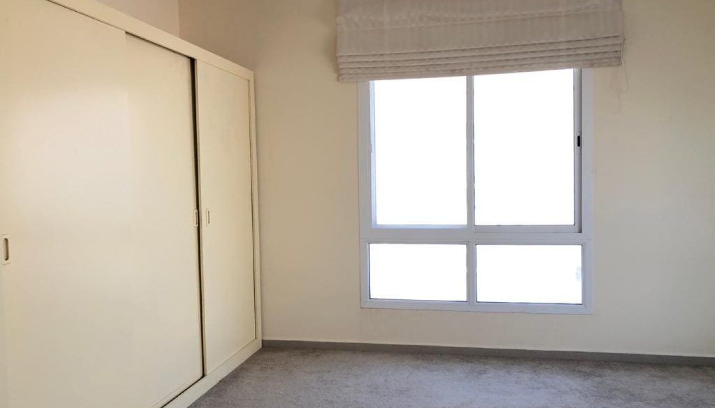 A bright, empty room with beige walls, a light gray carpet, a large window with a white curtain, and a closed white wardrobe.