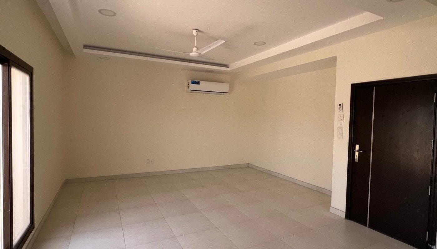 An empty room in a Sanad villa awaits, featuring tiled flooring, a ceiling fan, mounted air conditioning unit, large window, and a dark wooden door. Perfect for rent!