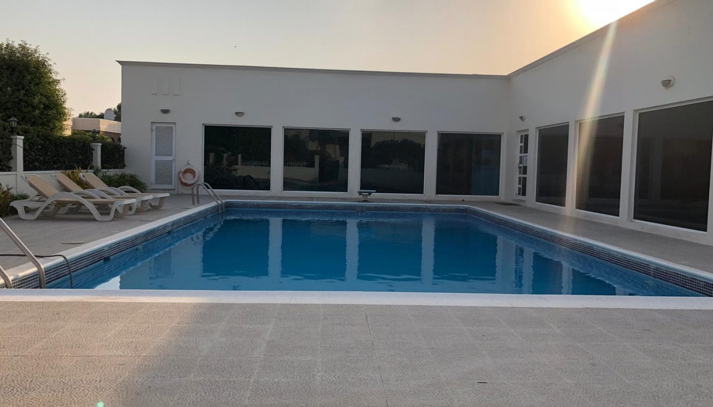 A rectangular swimming pool in front of a modern white 3 Bedrooms Villa with large windows and lounge chairs at sunset.
