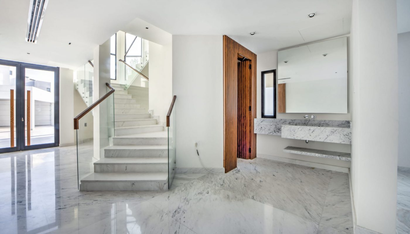 Modern interior with marble flooring, an open staircase with glass railing, a wall-mounted marble sink, large mirror, and floor-to-ceiling windows letting in natural light.