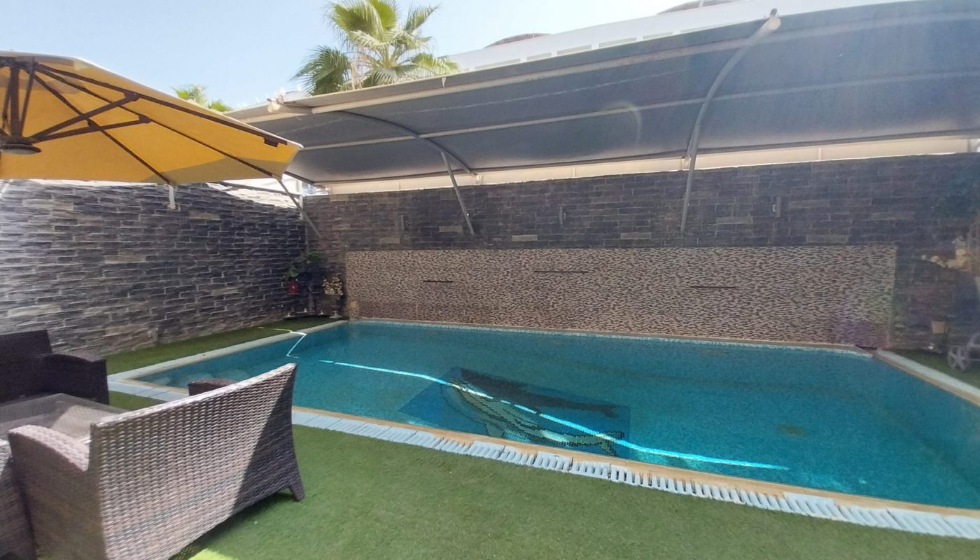 Rectangular outdoor swimming pool with clear water, shaded by a large canopy, next to a stone wall. Wicker chairs and a yellow patio umbrella are on artificial grass beside the pool.