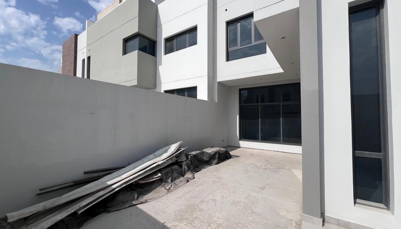 A modern white building with large windows and a small enclosed concrete yard; construction materials and debris are stacked along the left side.