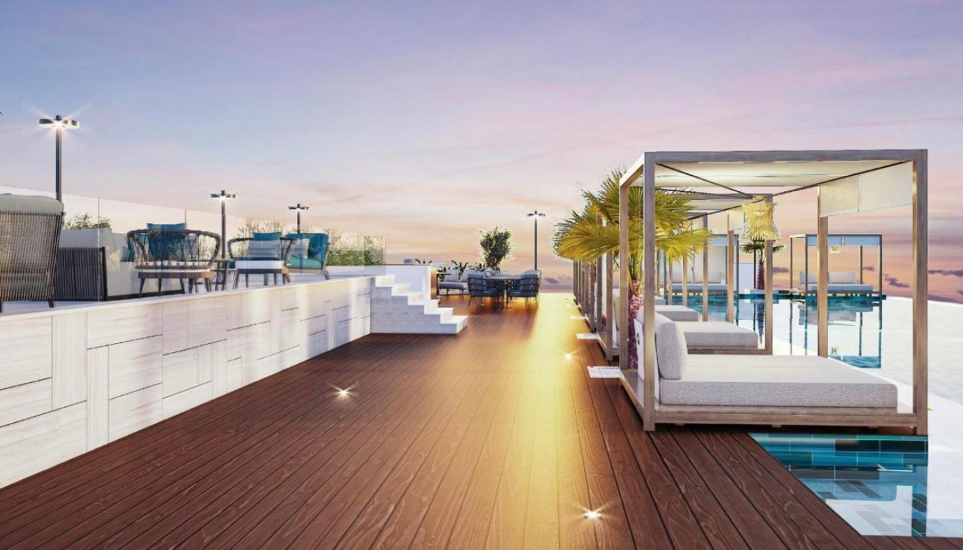 Rooftop deck with wooden flooring, modern lounge chairs, covered cabanas, potted palm trees, and a pool, under a colorful sunset sky.