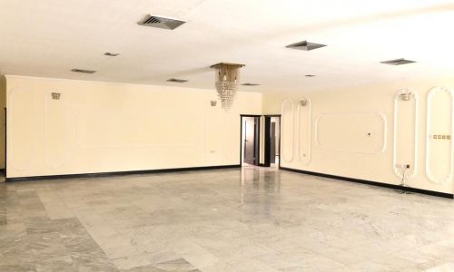 A spacious, empty room with marble flooring, light-colored walls, modern light fixtures, and a crystal chandelier hanging from the ceiling. Several doorways and vents are also visible.