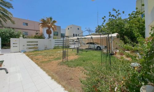 A backyard with a mix of grass and dirt areas, bordered by plants and shrubs. An auto draft white vehicle is parked under a canopy, and several modern houses are visible in the background.