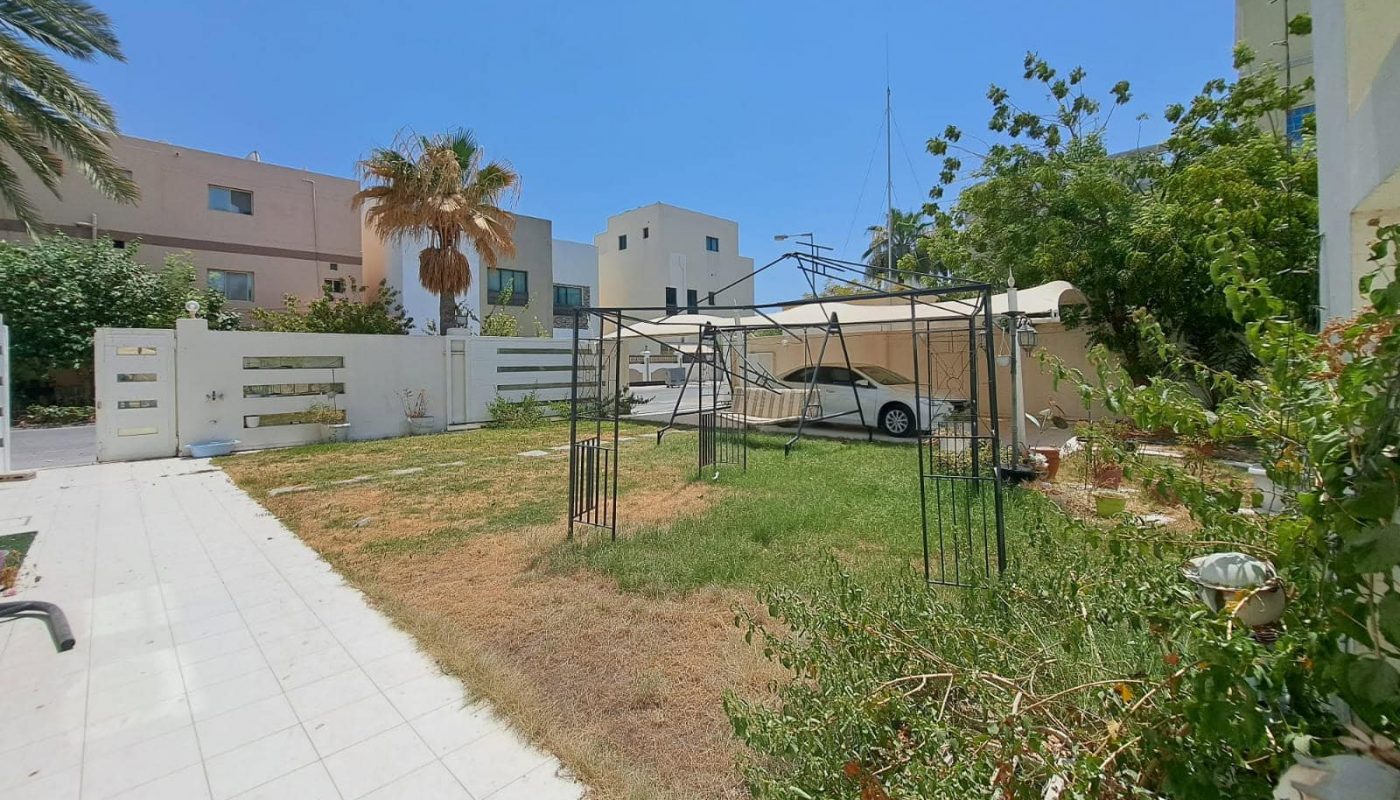 A backyard with a mix of grass and dirt areas, bordered by plants and shrubs. An auto draft white vehicle is parked under a canopy, and several modern houses are visible in the background.