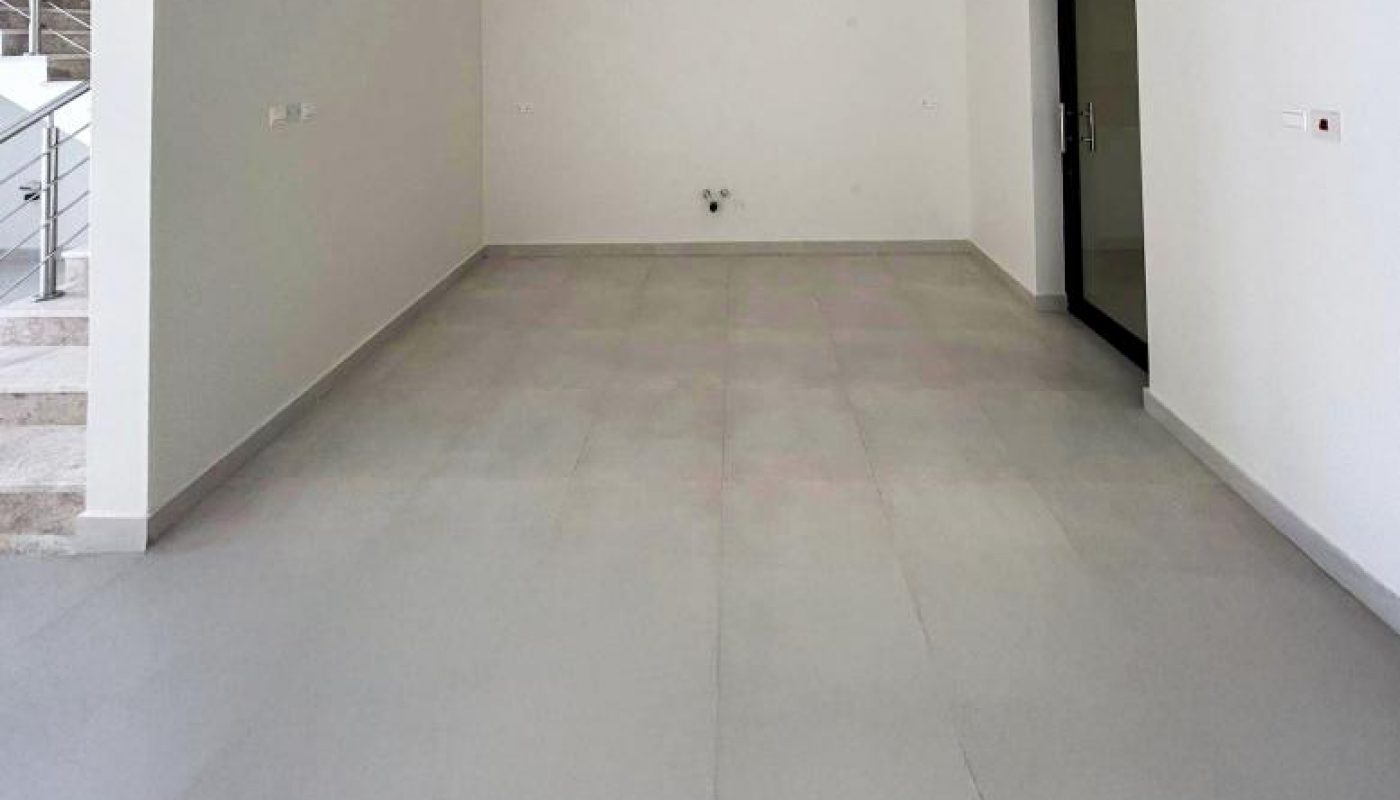 Empty room with light gray tiled floor, white walls, and recessed ceiling lights. A black door is on the right wall; stairs are visible to the left.