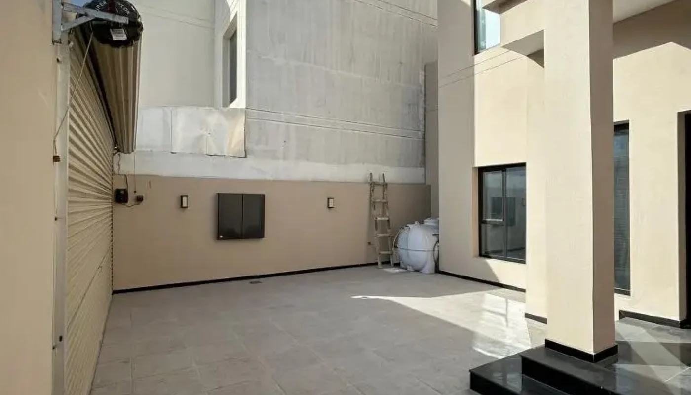 A modern outdoor courtyard with tiled flooring, beige walls, a closed garage door, ladder, and water tanks under a cloudy sky.