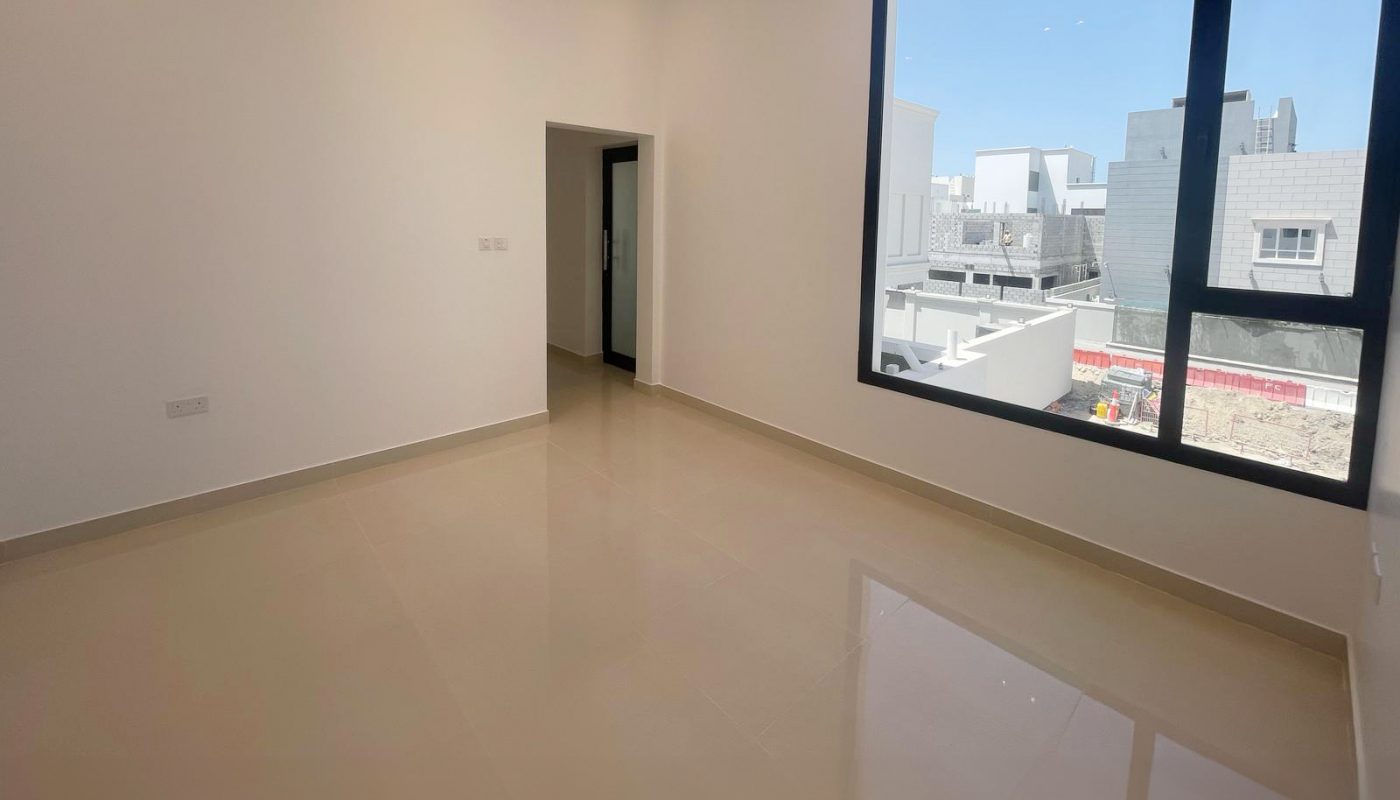 Empty, unfurnished room with cream-colored tile floors, white walls, recessed ceiling lights, and a large window overlooking modern buildings outside.