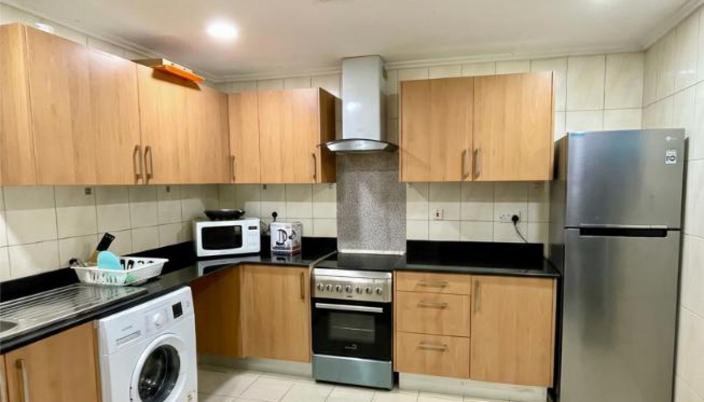 Modern kitchen with wooden cabinets, a silver refrigerator, washing machine, oven with stovetop, microwave, kettle, and tiled floor and walls.