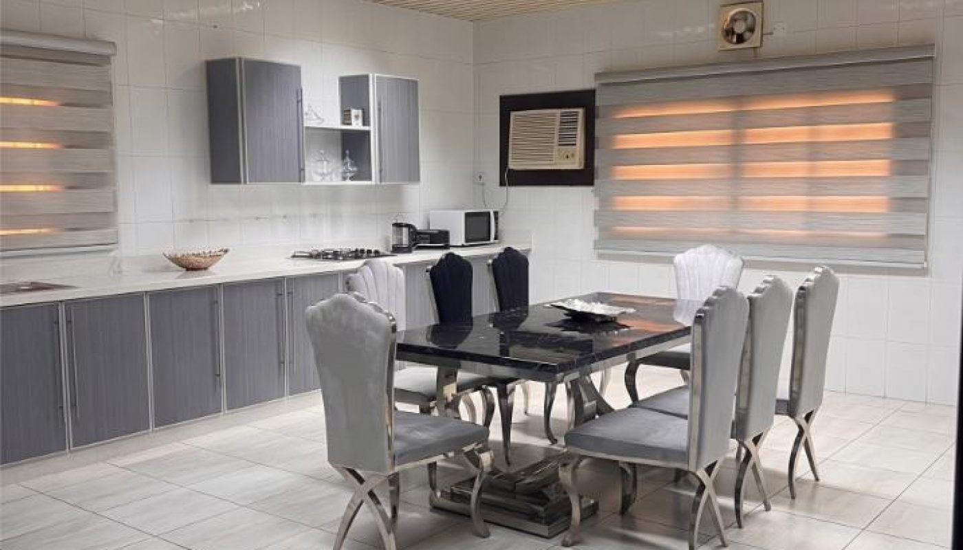 Modern kitchen and dining area with gray cabinets, a set table for six, appliances on the counter, tiled walls, and striped window blinds.