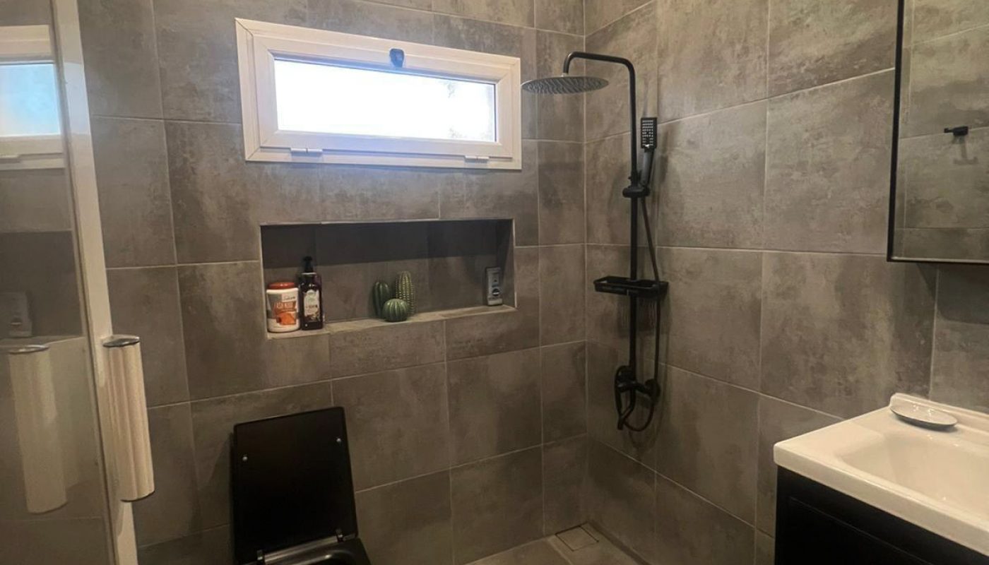 Modern bathroom with gray tiles, a black toilet, open shower with black fixtures, a rectangular sink, and a small window above a built-in shower shelf containing toiletries and a small plant.