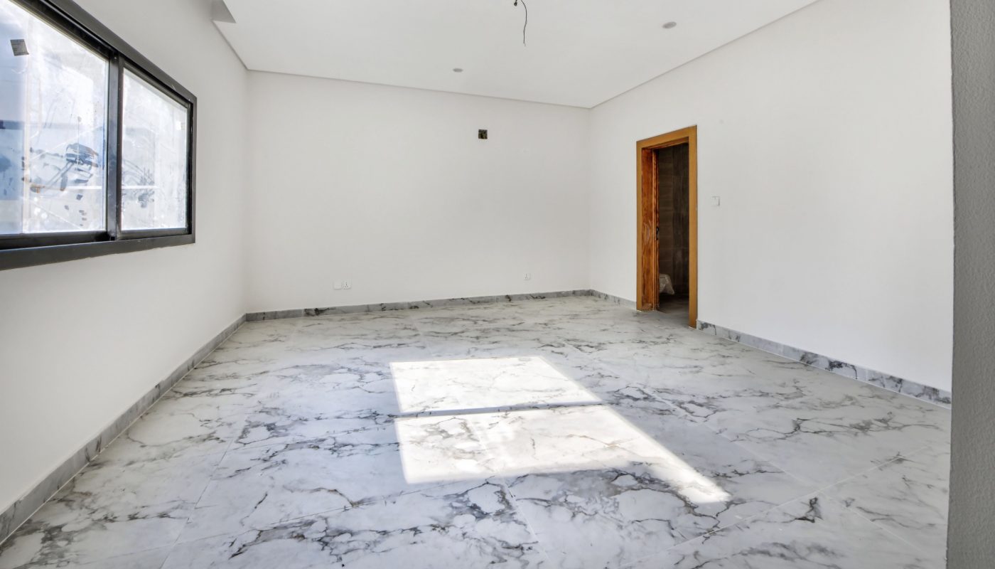 Empty room with white walls, large window, marble tile floor, and an open wooden door. Light from the window casts a shadow on the floor. Ceiling has recessed lighting fixtures.