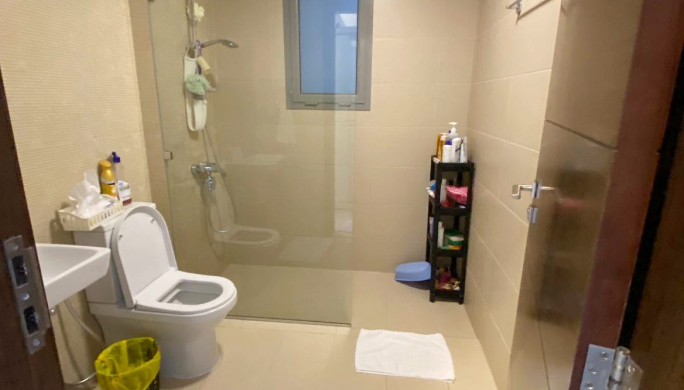 A bathroom with a toilet, sink, shower area separated by glass, window, wall-mounted shelf with toiletries, and two white floor mats.