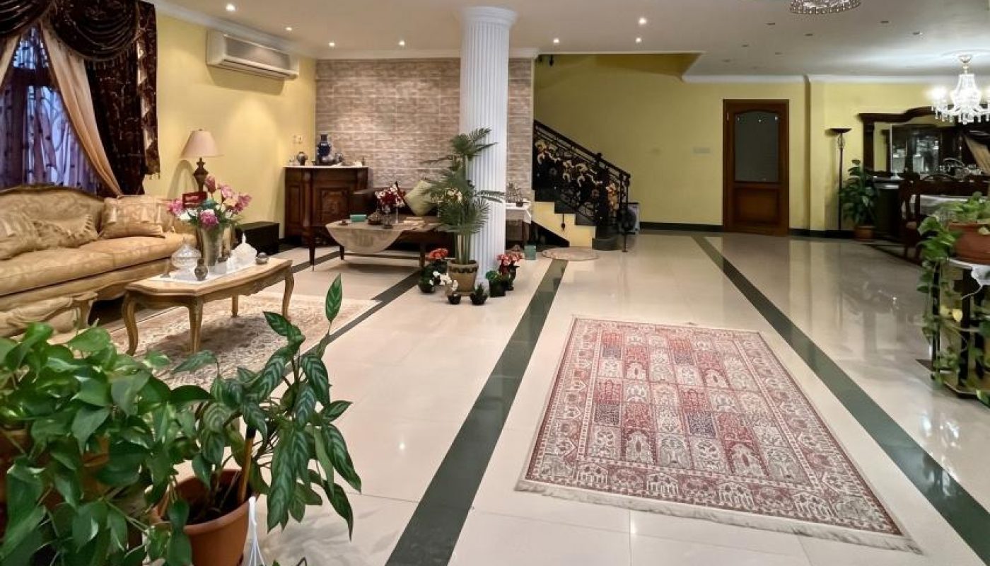 Spacious living room with cream-colored sofas, potted plants, a patterned rug, a decorative column, staircase with ornate railing, and glossy tiled floor.
