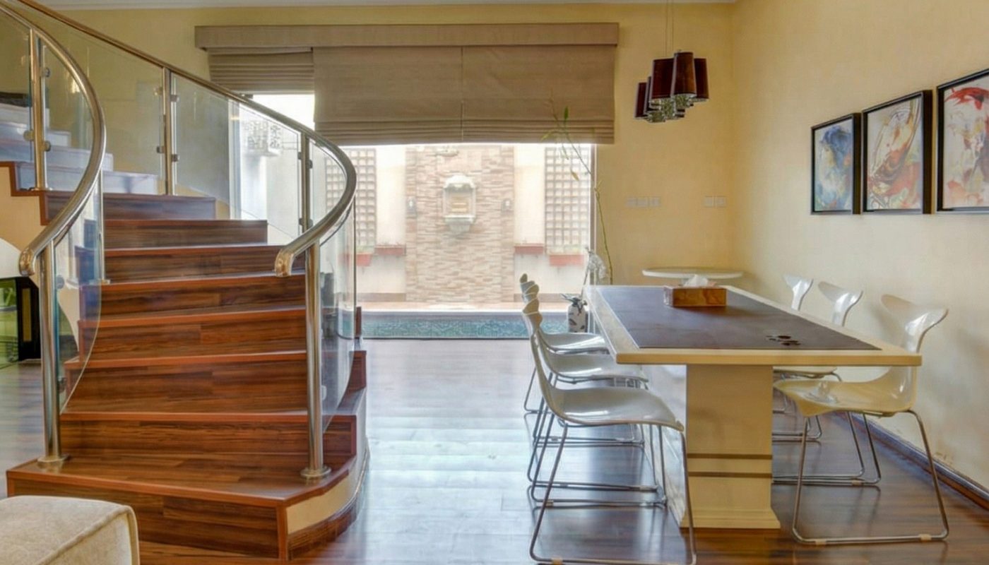 Modern dining area with a rectangular table, six white chairs, wooden flooring, and a curved staircase with glass railing in the background.
