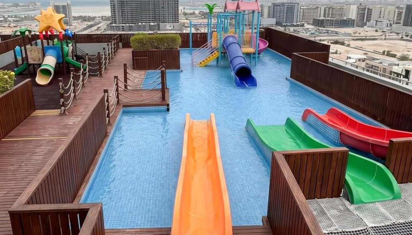 Rooftop pool area with multiple colorful water slides and play structures, surrounded by wooden decking, overlooking a cityscape under a cloudy sky.