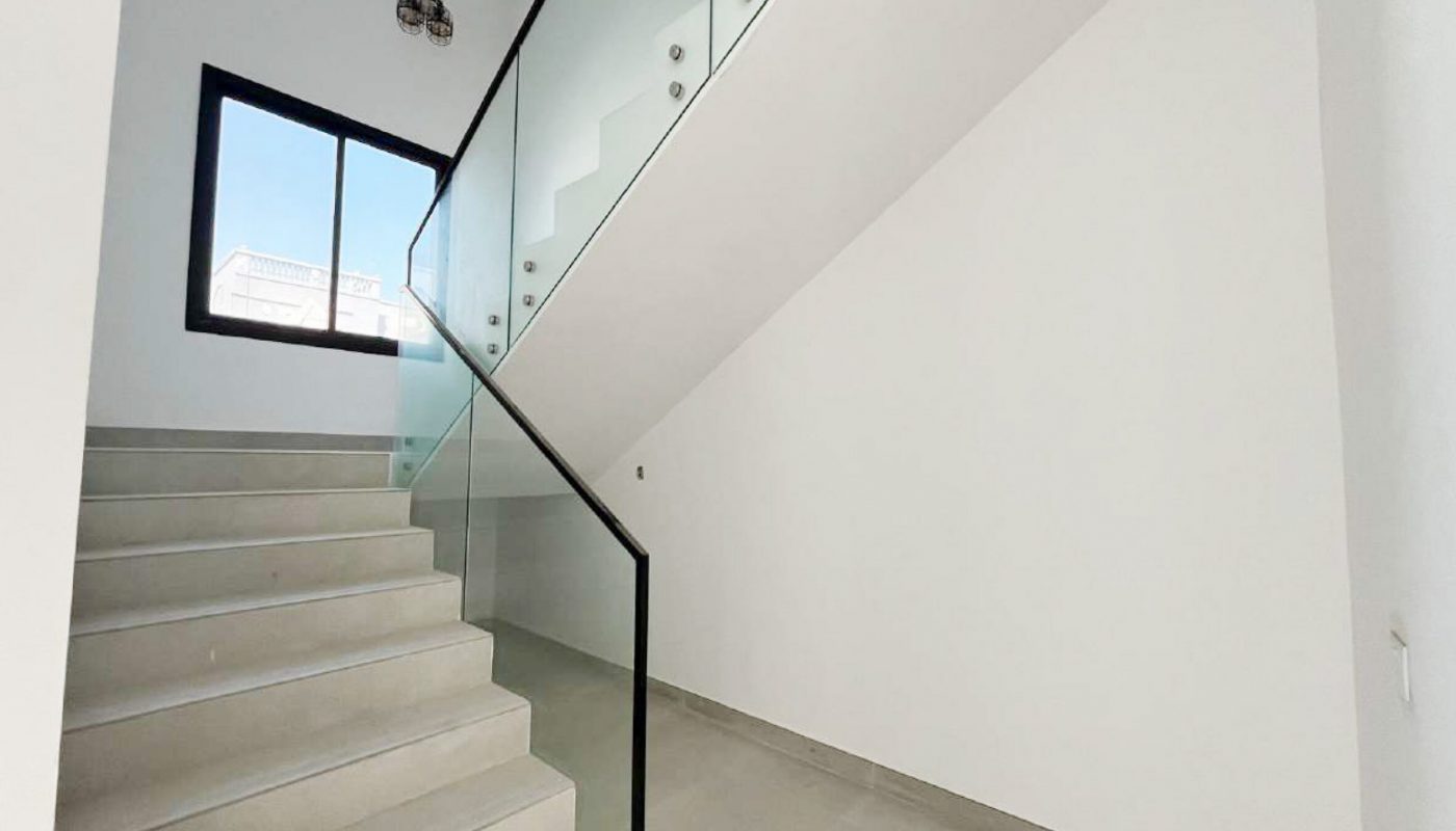 A modern staircase with glass railing, light grey steps, a large window, and white walls in a minimalistic interior space.