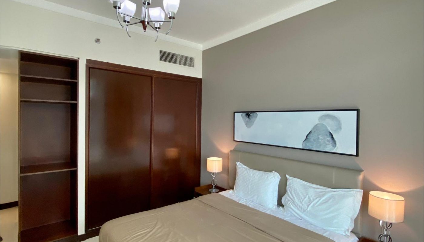 A neatly made bed with beige bedding in a modern bedroom, featuring two nightstands with lamps, a large wardrobe, and abstract wall art above the headboard.