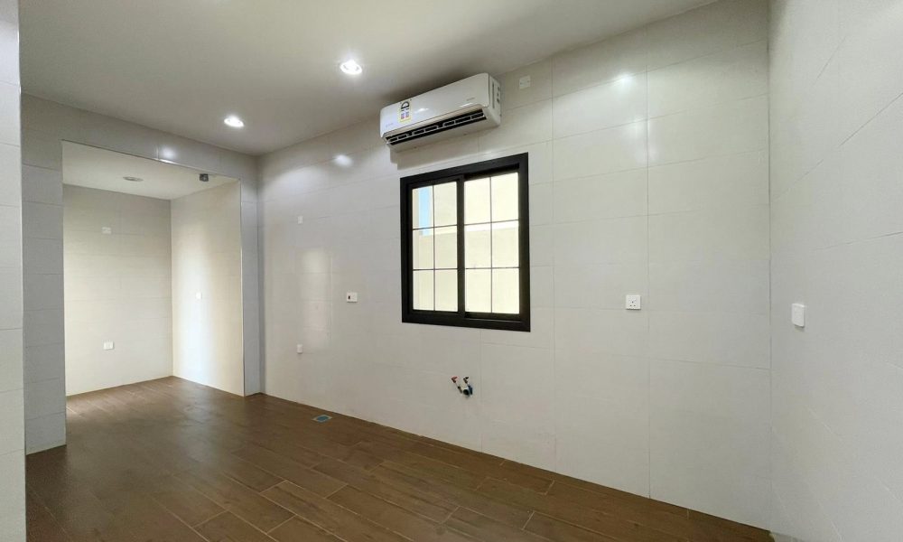 Empty room with wooden floor, gray walls, ceiling lights, a black-framed window, and a wall-mounted air conditioner. In the background, another unfurnished room is visible. This villa in Diyar Al Muharraq is for sale and offers a spacious layout perfect for customization.