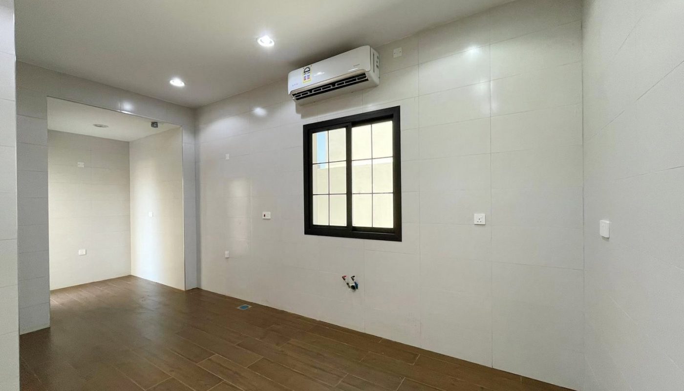Empty room with wooden floor, gray walls, ceiling lights, a black-framed window, and a wall-mounted air conditioner. In the background, another unfurnished room is visible. This villa in Diyar Al Muharraq is for sale and offers a spacious layout perfect for customization.