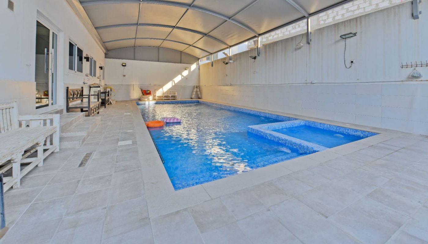An indoor swimming pool with a covered ceiling, surrounded by white tiles, benches, and a small table.