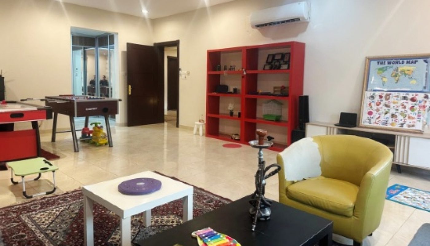 A playroom with games tables, children’s toys, bookshelves, rugs, a world map on the wall, and various tables and chairs arranged throughout the space.